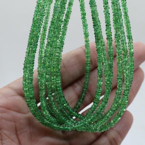 May include: A close-up of multiple strands of green gemstone beads. The beads are small, square-cut, and a vibrant green colour. The strands are held in a person's hand against a white background. The beads appear to be strung together to create a necklace.