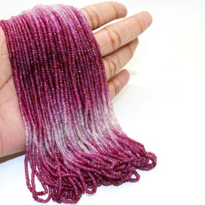 May include: Strands of small, faceted beads in shades of pink and purple are held in a hand against a white background. The beads transition from a deep magenta to a lighter, almost white color. The beads are likely gemstones.