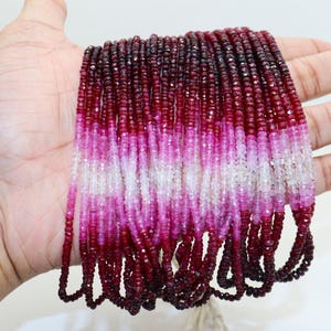 May include: Strands of small, faceted beads in a gradient of colors from deep red to light pink and white. The beads are held in a hand, showcasing the color variations and the texture of the gemstones. The beads are likely for jewelry making.