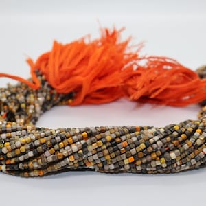 Op de afbeelding: Een streng kleine, vierkante edelsteen kralen in tinten van bruin, grijs, wit en oranje. De kralen zijn geregen aan een koord en hebben een kwast aan het einde.