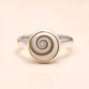 Könnte beinhalten: Ein silberner Ring mit einem kreisförmigen, cremefarbenen Stein mit einem Spiralmuster. Der Stein ist in einer goldfarbenen Lünette gefasst, und das Ringband ist silberfarben. Der Ring ist ein Schmuckstück.