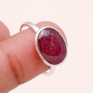 May include: A silver ring featuring a large, oval-cut ruby gemstone. The ruby is a deep, rich red color and is set in a simple, polished silver band. The ring is shown on a finger.