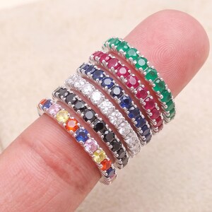 Peut inclure: Une pile de sept bagues éternité fines en argent, chacune ornée d'une rangée de pierres précieuses rondes de couleurs distinctes, notamment vert, rouge, bleu foncé, blanc transparent, noir et une bande multicolore avec des pierres orange, bleu, rose et jaune.