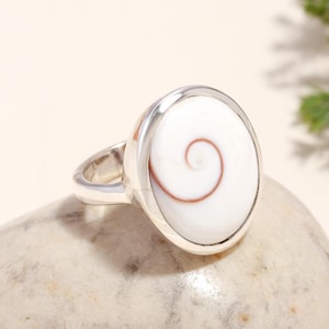 Può includere: Un anello in argento con un inserto ovale in conchiglia occhio di Shiva bianca. La conchiglia presenta un motivo a spirale naturale color marrone rossastro centrato su una superficie bianca liscia e lucida, incastonato in una fascia d'argento.