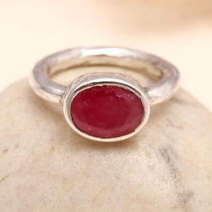 Op de afbeelding: Een zilveren ring met een grote, ovaal geslepen robijn edelsteen. De ring heeft een getextureerde band en een bezel setting die de levendige rode steen vasthoudt. De ring wordt gepresenteerd op een lichtgekleurd, getextureerd oppervlak.