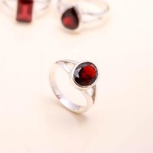 May include: A silver ring featuring a large, oval, deep red gemstone. The band splits into two parts either side of the gemstone. Other rings with red gemstones are visible in the background.