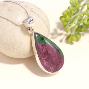 Può includere: Un ciondolo a goccia in rubino zoisite incastonato in una montatura in argento lucido, sospeso a una sottile catena a serpente in argento. La gemma presenta un centro magenta intenso con vivaci inclusioni minerali verdi sulla superficie.