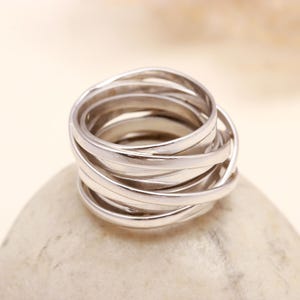 May include: A silver ring featuring multiple interwoven bands, forming a layered design. The ring is presented on a light-toned, textured surface, highlighting its detailed construction and metallic lustre. This piece of jewellery is suitable for everyday wear.