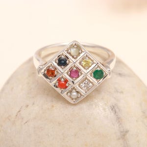 May include: A silver ring featuring a diamond-shaped face with a grid of nine small, round, multi-colored gemstones. The stones include a white pearl, a dark blue sapphire, a yellow sapphire, a red ruby, a green emerald, and orange coral.