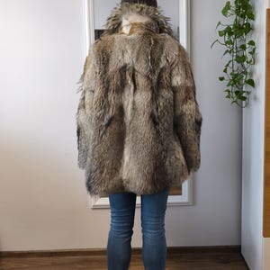 K&ouml;nnte beinhalten: Ein brauner und grauer Pelzmantel mit strukturierter Oberfl&auml;che. Der Mantel hat einen breiten Kragen und wird mit blauen Jeans und schwarzen Stiefeln getragen. Die Person steht vor einem Spiegel.