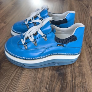 Puede incluir: Un par de zapatos de cuero azul con cordones blancos y una suela gruesa y en capas. Los zapatos tienen una puntera redondeada y un cuello acolchado. Las suelas tienen aproximadamente 5 cm de grosor.