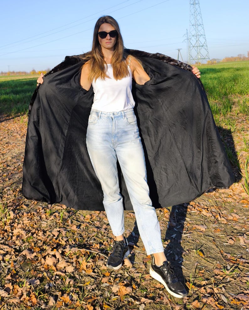 Puede incluir: Un abrigo largo negro con cuello de piel sint&eacute;tica est&aacute; abierto, revelando una camiseta sin mangas blanca y vaqueros azul claro. El abrigo se sostiene abierto, mostrando su forro oscuro. La persona lleva zapatillas negras y gafas de sol, de pie en un campo.