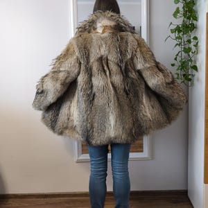 K&ouml;nnte beinhalten: Ein Vintage-Pelzmantel in Braun- und Graut&ouml;nen. Der Mantel hat eine weite, ausgestellte Form und ein strukturiertes Aussehen. Das Model tr&auml;gt blaue Jeans und schwarze Stiefel. Der Mantel ist der Blickfang, mit einem Spiegel und einer Wand im Hintergrund.