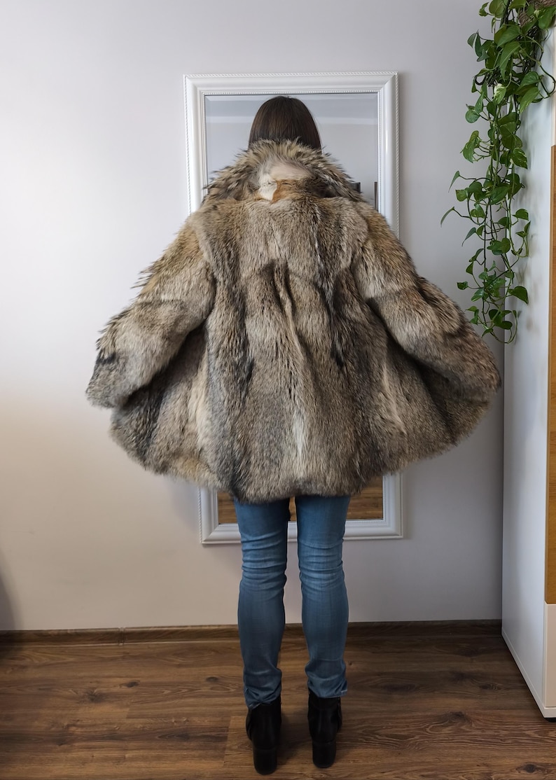 K&ouml;nnte beinhalten: Ein Vintage-Pelzmantel in Braun- und Graut&ouml;nen. Der Mantel hat eine weite, ausgestellte Form und ein strukturiertes Aussehen. Das Model tr&auml;gt blaue Jeans und schwarze Stiefel. Der Mantel ist der Blickfang, mit einem Spiegel und einer Wand im Hintergrund.