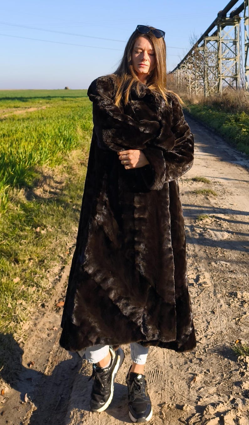 Puede incluir: Un abrigo largo de piel sint&eacute;tica marr&oacute;n oscuro. El abrigo es largo y tiene un aspecto afelpado y texturizado. La persona lleva zapatillas negras y vaqueros claros. El fondo muestra un camino de tierra y un campo verde.