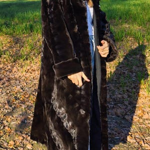 Puede incluir: Abrigo largo de piel sint&eacute;tica marr&oacute;n oscuro con cuello ancho y pu&ntilde;os. El abrigo se lleva sobre una camisa blanca y unos vaqueros azules. La persona lleva gafas de sol negras y zapatillas deportivas blancas y negras. El fondo es un campo.