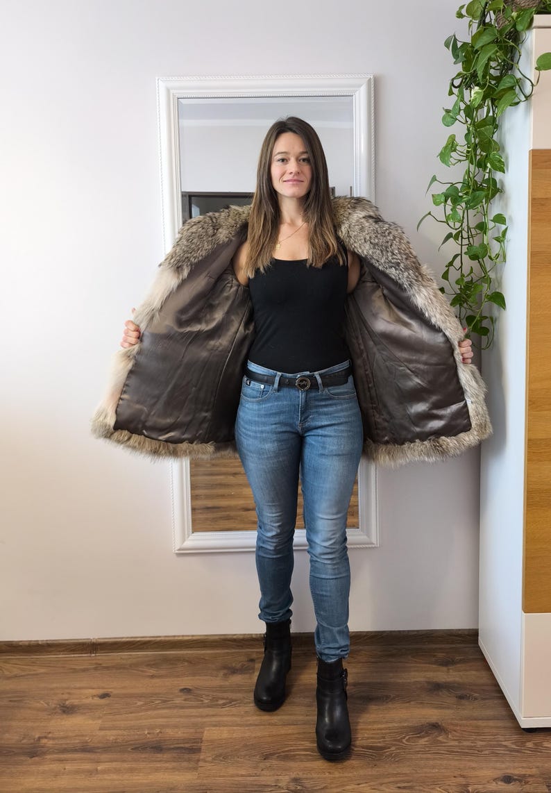 K&ouml;nnte beinhalten: Ein Mantel aus Kunstfell mit braunem Satinfutter, getragen mit einem schwarzen Top, blauen Jeans und schwarzen Stiefeln. Der Mantel ist offen und zeigt das Futter. Das Outfit wird durch einen schwarzen G&uuml;rtel erg&auml;nzt.