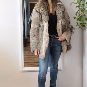 K&ouml;nnte beinhalten: Eine Person tr&auml;gt einen mehrfarbigen Pelzmantel in Braun-, Grau- und Cremet&ouml;nen. Sie tr&auml;gt au&szlig;erdem ein schwarzes Oberteil, eine blaue Jeans, einen schwarzen G&uuml;rtel und schwarze Stiefel. Der Hintergrund umfasst einen Spiegel und einen Holzboden.
