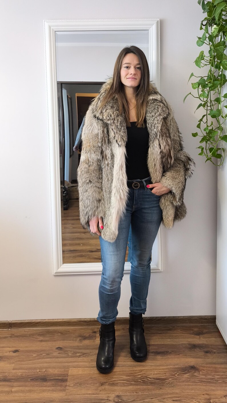K&ouml;nnte beinhalten: Eine Person tr&auml;gt einen mehrfarbigen Pelzmantel in Braun-, Grau- und Cremet&ouml;nen. Sie tr&auml;gt au&szlig;erdem ein schwarzes Oberteil, eine blaue Jeans, einen schwarzen G&uuml;rtel und schwarze Stiefel. Der Hintergrund umfasst einen Spiegel und einen Holzboden.