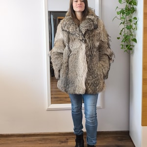 K&ouml;nnte beinhalten: Ganzk&ouml;rperaufnahme einer Person in einem Kunstfellmantel. Der Mantel hat eine Mischung aus Grau- und Braunt&ouml;nen und einen gro&szlig;en Kragen. Die Person tr&auml;gt eine blaue Jeans und schwarze Stiefel. Ein Spiegel und Gr&uuml;nzeug sind im Hintergrund.