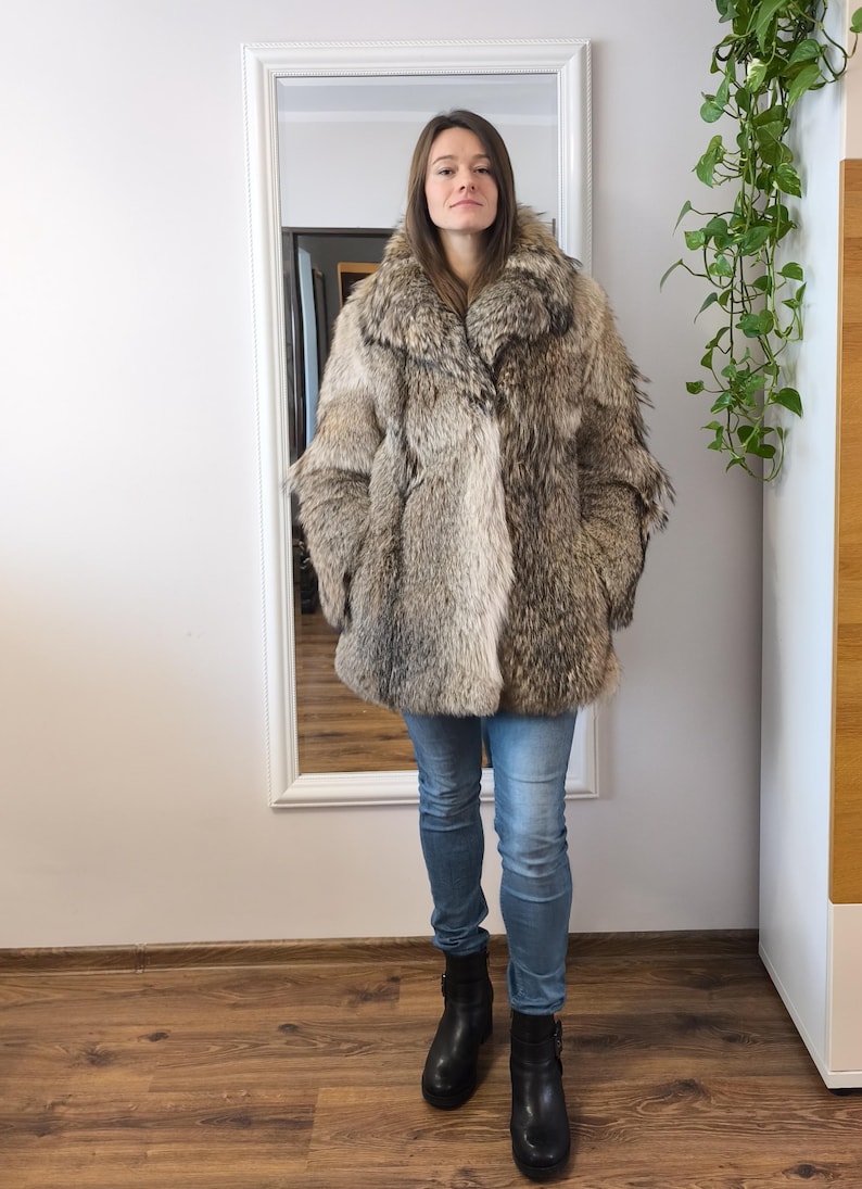 K&ouml;nnte beinhalten: Ganzk&ouml;rperaufnahme einer Person in einem Kunstfellmantel. Der Mantel hat eine Mischung aus Grau- und Braunt&ouml;nen und einen gro&szlig;en Kragen. Die Person tr&auml;gt eine blaue Jeans und schwarze Stiefel. Ein Spiegel und Gr&uuml;nzeug sind im Hintergrund.