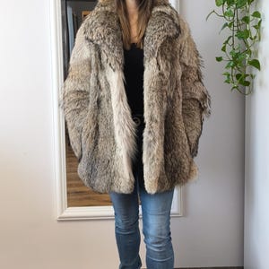 K&ouml;nnte beinhalten: Eine Frau in einem &uuml;bergro&szlig;en Pelzmantel in Braun-, Beige- und Graut&ouml;nen. Sie tr&auml;gt auch ein schwarzes Oberteil, blaue Jeans und schwarze Stiefel. Der Mantel hat einen breiten Kragen und eine lockere Passform und wirkt luxuri&ouml;s.