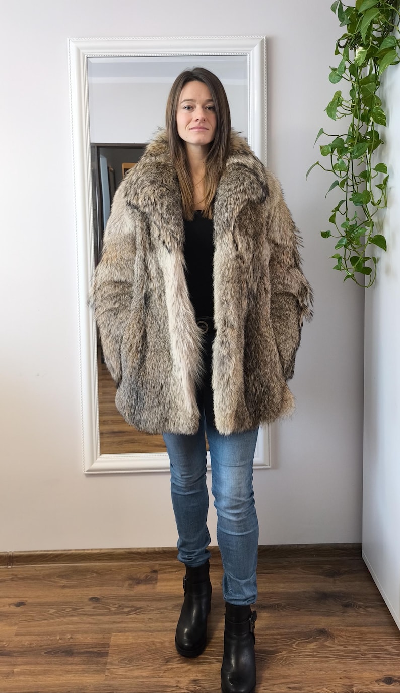 K&ouml;nnte beinhalten: Eine Frau in einem &uuml;bergro&szlig;en Pelzmantel in Braun-, Beige- und Graut&ouml;nen. Sie tr&auml;gt auch ein schwarzes Oberteil, blaue Jeans und schwarze Stiefel. Der Mantel hat einen breiten Kragen und eine lockere Passform und wirkt luxuri&ouml;s.