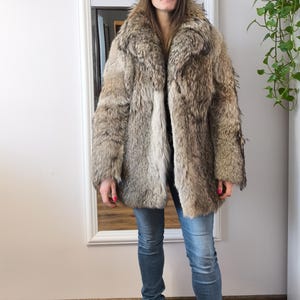 K&ouml;nnte beinhalten: Ein Kunstfellmantel in Braun- und Graut&ouml;nen mit breitem Kragen und langen &Auml;rmeln. Der Mantel wird mit blauen Jeans und schwarzen Stiefeletten getragen. Die Person steht vor einem wei&szlig; gerahmten Spiegel.