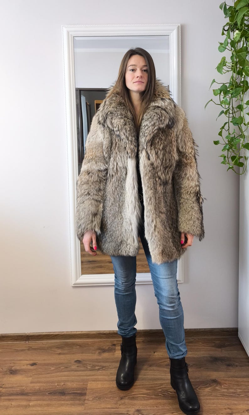 K&ouml;nnte beinhalten: Ein Kunstfellmantel in Braun- und Graut&ouml;nen mit breitem Kragen und langen &Auml;rmeln. Der Mantel wird mit blauen Jeans und schwarzen Stiefeletten getragen. Die Person steht vor einem wei&szlig; gerahmten Spiegel.