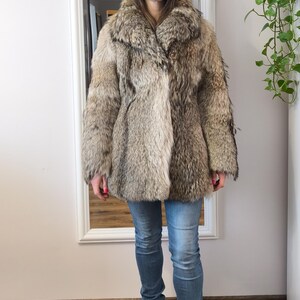 K&ouml;nnte beinhalten: Eine Person tr&auml;gt einen Vintage-Pelzmantel. Der Mantel hat eine Mischung aus Braun- und Graut&ouml;nen, einen gro&szlig;en Kragen und einen Knopfverschluss. Sie tr&auml;gt auch blaue Jeans und schwarze Stiefel. Der Hintergrund umfasst einen wei&szlig; gerahmten Spiegel und einen Holzboden.