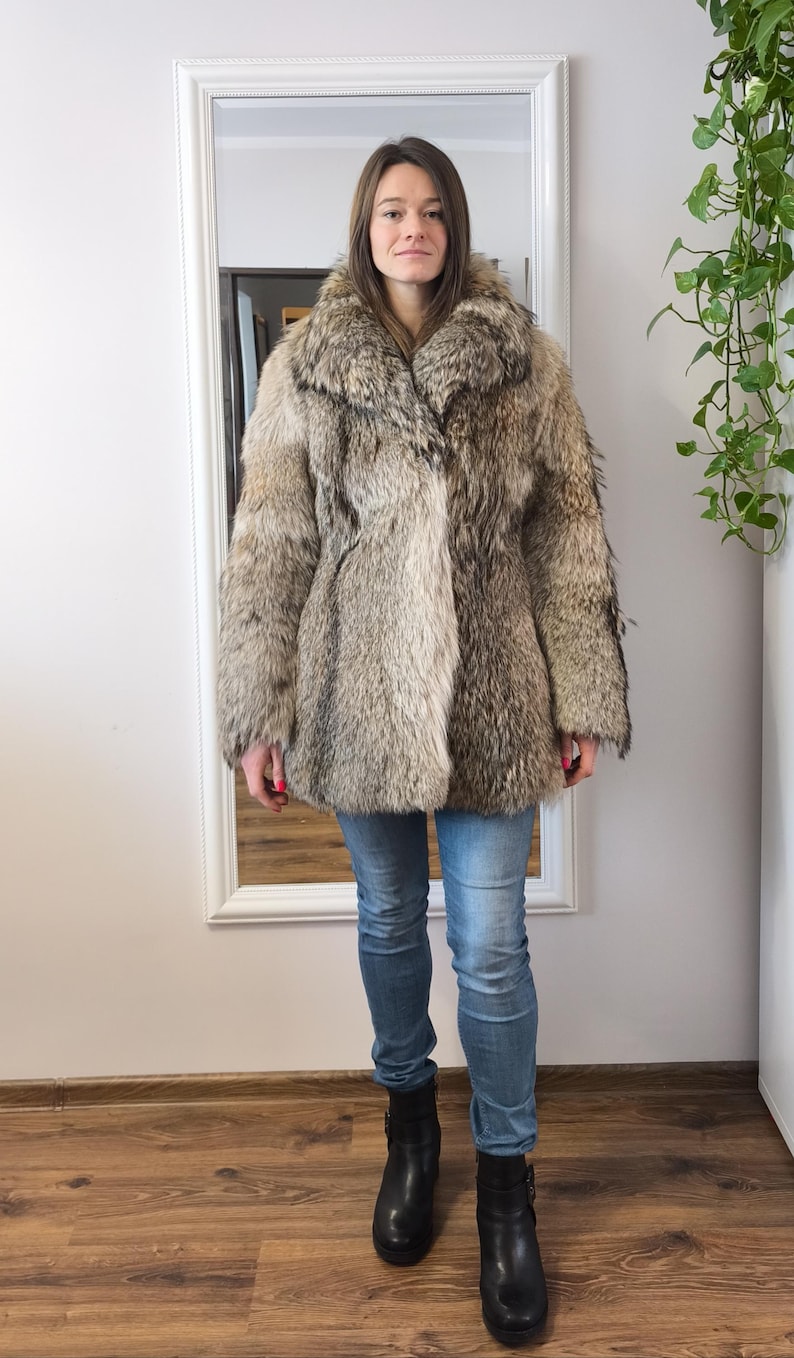 K&ouml;nnte beinhalten: Eine Person tr&auml;gt einen Vintage-Pelzmantel. Der Mantel hat eine Mischung aus Braun- und Graut&ouml;nen, einen gro&szlig;en Kragen und einen Knopfverschluss. Sie tr&auml;gt auch blaue Jeans und schwarze Stiefel. Der Hintergrund umfasst einen wei&szlig; gerahmten Spiegel und einen Holzboden.