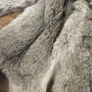 K&ouml;nnte beinhalten: Nahaufnahme eines luxuri&ouml;sen Kunstfelltextils in Grau-, Braun- und Wei&szlig;t&ouml;nen. Das weiche, strukturierte Material ist in Falten angeordnet und zeigt das detaillierte Muster und die verschiedenen Farbt&ouml;ne. Das Fell wirkt weich und einladend, perfekt f&uuml;r die Inneneinrichtung.