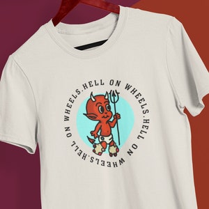 May include: Off-white t-shirt featuring a cartoon devil with a trident and roller skates inside a blue circle. The text "HELL ON WHEELS" is printed around the circle in black. The shirt is hanging on a wooden hanger.