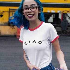 May include: White t-shirt with red trim and the word "CIAO" in black letters. The person is wearing blue denim shorts and a red and blue plaid shirt. The person is also wearing black glasses.