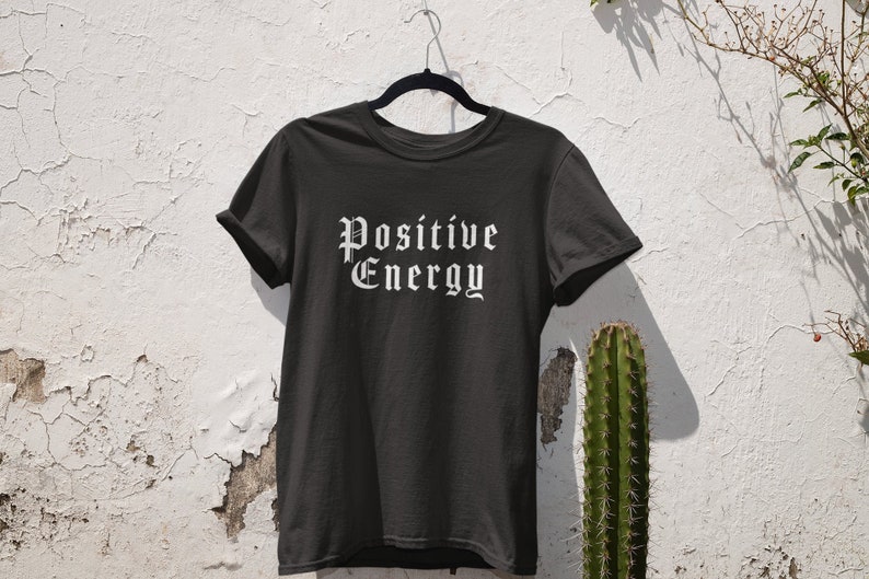 May include: Black t-shirt with the words "POSITIVE ENERGY" in a white gothic font. The shirt is hanging on a black hanger against a textured white wall. A cactus and plant are visible on the right side.