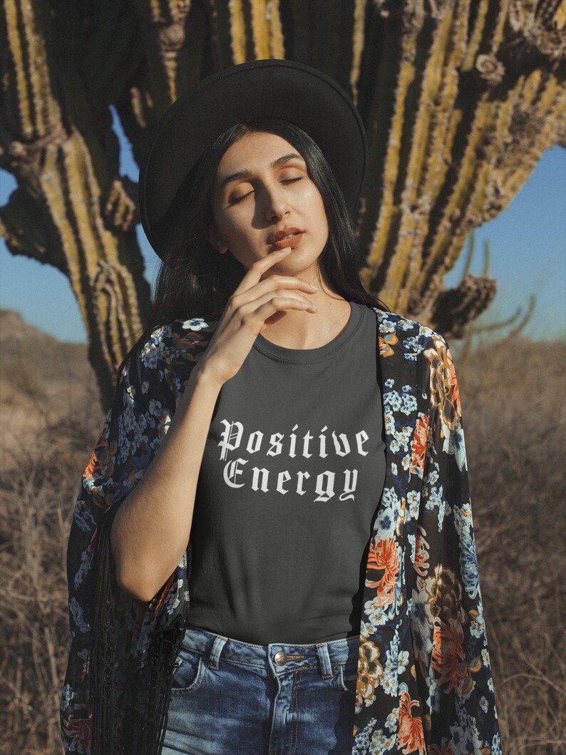 May include: A dark gray t-shirt with the words "Positive Energy" in white gothic font. The person is wearing a black hat, blue jeans, and a floral patterned kimono. The background features a large cactus and a clear blue sky.