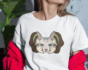 May include: White t-shirt featuring a graphic of a cat's face split into two halves, each side resembling a woman's face. The cat's face is light beige with brown hair and green eyes. The woman is wearing a red jacket and blue and white striped jeans.