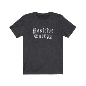 May include: Dark gray t-shirt with the words "Positive Energy" in a white gothic font. The shirt has short sleeves and a crew neck. The fabric appears to be a soft, heathered material.