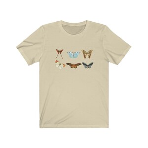 May include: A beige t-shirt featuring six colorful butterfly illustrations. The butterflies are in various colors, including red, blue, brown, and white. The shirt is made of a soft material and has short sleeves.