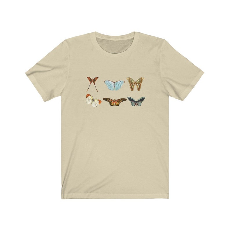May include: A beige t-shirt featuring six colorful butterfly illustrations. The butterflies are in various colors, including red, blue, brown, and white. The shirt is made of a soft material and has short sleeves.