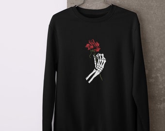 May include: Black crew neck sweatshirt with a graphic of a skeleton hand holding a red rose. The design is centrally placed. The sweatshirt is suspended on a metal hanger against a neutral backdrop.