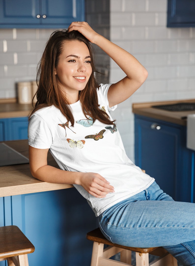 May include: White t-shirt featuring a collection of colorful butterfly illustrations. The shirt is worn by a person in blue jeans, seated in a kitchen setting with blue cabinets and a wooden countertop.