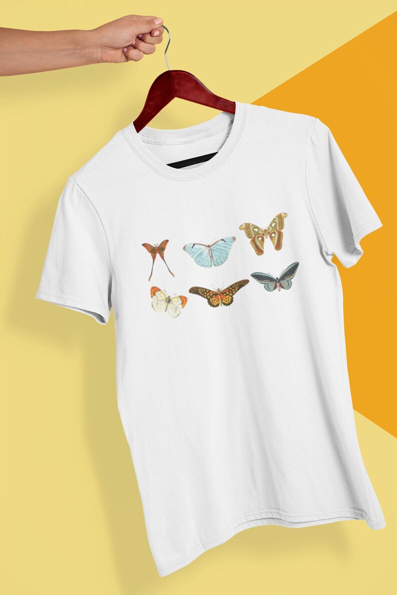 May include: White t-shirt featuring a collection of colorful butterfly illustrations. The butterflies vary in size and color, with details like orange, blue, and brown wings. The shirt is displayed on a wooden hanger against a yellow background.
