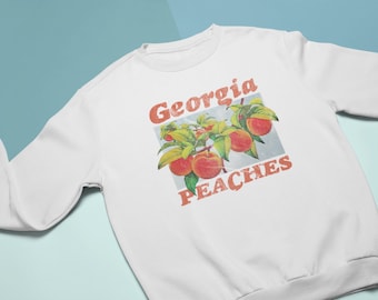 Könnte beinhalten: Weißes Sweatshirt mit einem Grafikdesign von Pfirsichen an einem Ast und dem Schriftzug "Georgia PEACHES" in Orange. Das Design hat einen Vintage-Look. Das Sweatshirt ist aus weichem Material.