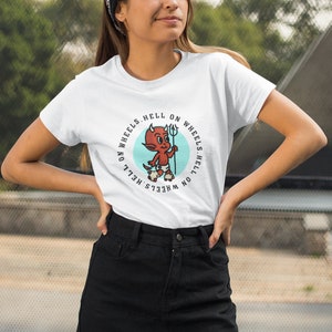 May include: White t-shirt featuring a cartoon devil with a trident, roller skates, and the text "HELL ON WHEELS." The design is in a circular format with a light blue background. The shirt is worn by a person wearing a black bandana and a black skirt.