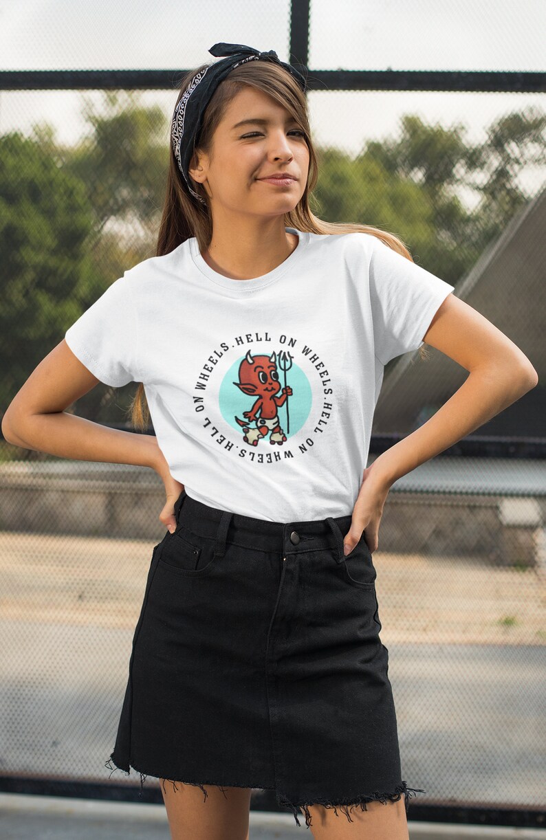 May include: White t-shirt featuring a cartoon devil with a trident, roller skates, and the text "HELL ON WHEELS." The design is in a circular format with a light blue background. The shirt is worn by a person wearing a black bandana and a black skirt.