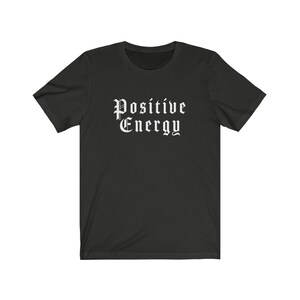 May include: A black t-shirt with the words "Positive Energy" printed in a gothic font. The text is in white and centered on the front of the shirt. The shirt has short sleeves and a crew neck.