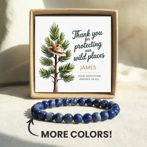 May include: A blue beaded bracelet sits in front of a gift box containing a card. The card features a tree with a hat and the text "Thank you for protecting our wild places" and "JAMES". The words "MORE COLORS!" are also visible.