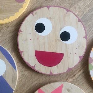 Könnte beinhalten: Ein rundes Holzbrett, das mit einem lächelnden Gesicht bemalt ist. Das Gesicht hat große weiße Augen mit schwarzen Pupillen, einen roten Mund und rosa Sommersprossen. Das Brett ist hellbraun mit einem rosa Rand.