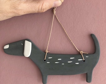 May include: A gray ceramic dachshund ornament with white markings hanging from a brown string. The ornament is a simple, stylized design of a dachshund with a long body and short legs.